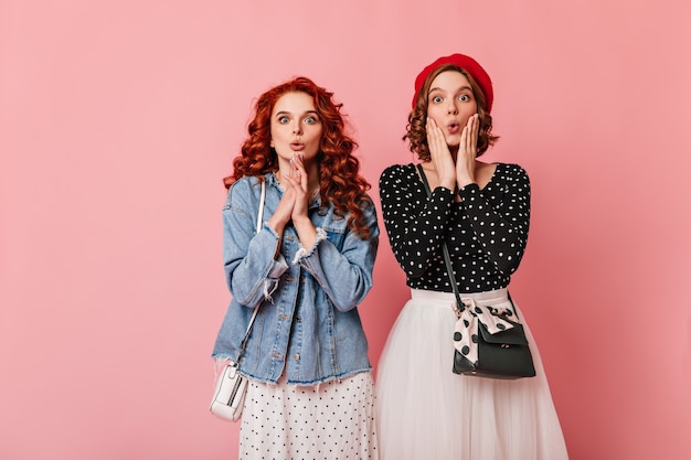 Mujer de jengibre sorprendida posando con un amigo. Foto de estudio de dos niñas expresando asombro sobre fondo rosa.