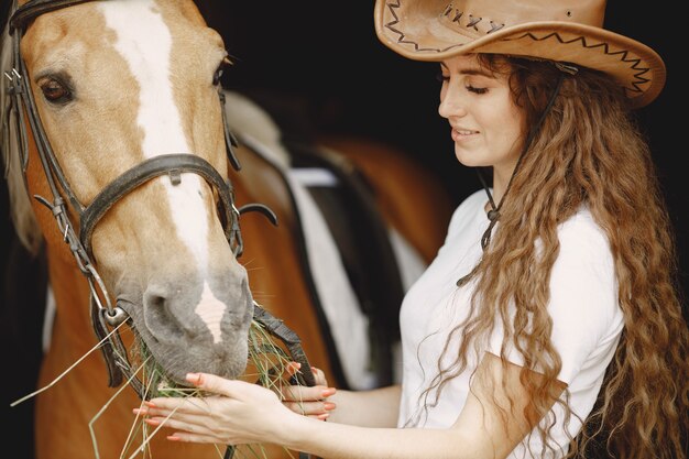 Mujer jinete alimentando a su caballo con heno en un establo. La mujer tiene el pelo largo y una camiseta blanca. El fondo es oscuro y negro.