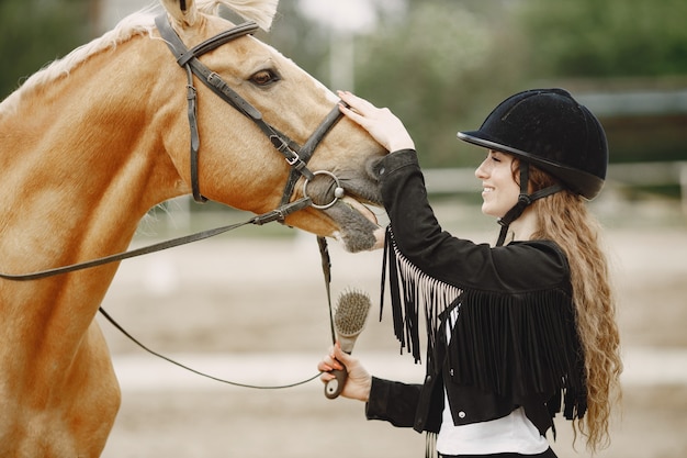 Mujer jinete hablando con su caballo en un rancho. La mujer tiene cabello largo y ropa negra. Ecuestre femenina tocando su caballo marrón.