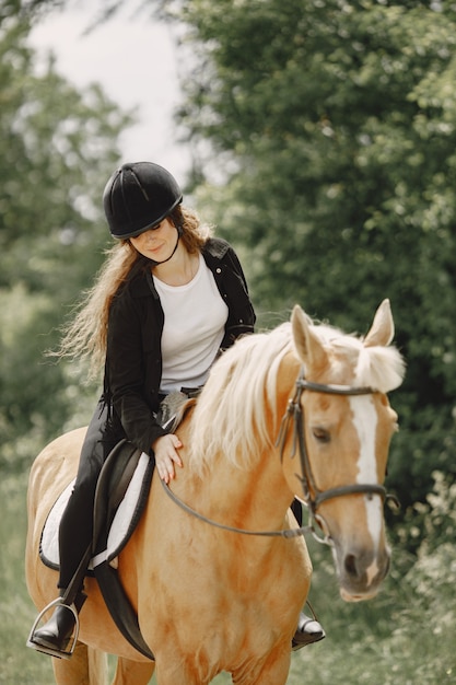 Mujer jinete montando su caballo en un rancho. La mujer tiene cabello largo y ropa negra. Ecuestre femenina tocando su caballo marrón.