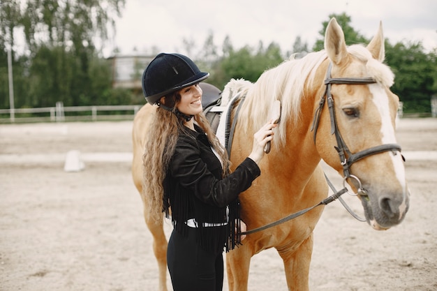 Mujer jinete peina su caballo en un rancho. La mujer tiene cabello largo y ropa negra. Ecuestre femenina tocando su caballo marrón.