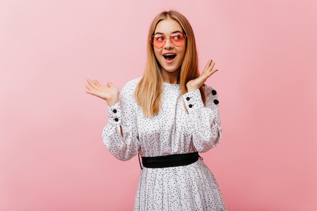 La mujer de Jocund usa gafas de sol rosas jugando durante la sesión de retratos de moda. Hermosa mujer caucásica en vestido blanco posando con una sonrisa sincera.