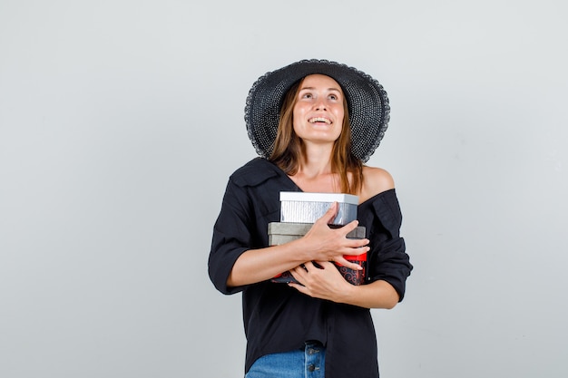 Mujer joven abrazando cajas de regalo mientras mira hacia arriba en camisa, pantalones cortos, sombrero y mirando feliz