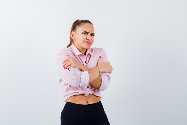 Mujer joven abrazándose a sí misma en camisa casual, pantalones y mirando fría