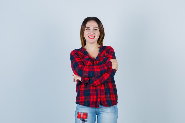 Mujer joven abrazándose a sí misma, sonriendo con camisa a cuadros, jeans y mirando alegre. vista frontal.