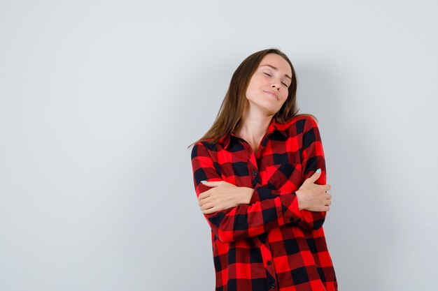 Mujer joven abrazándose a sí misma, sonriendo con camisa a cuadros y luciendo radiante. vista frontal.