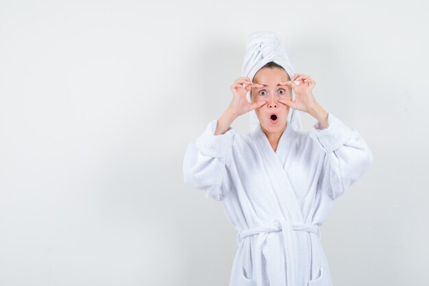 Mujer joven abriendo los ojos con los dedos en bata de baño blanca, toalla y mirando consternado. vista frontal.