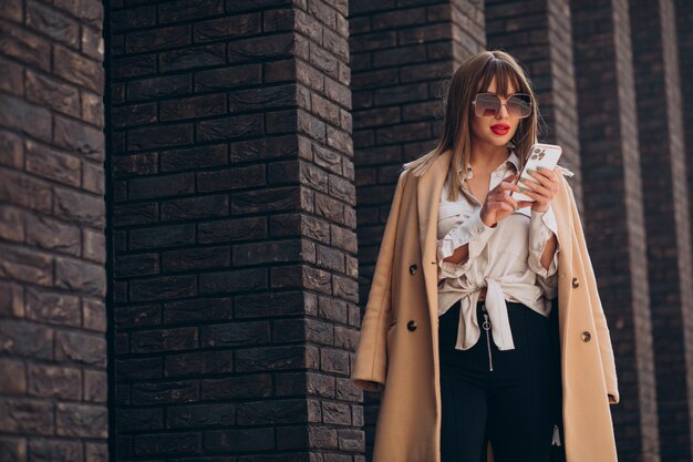 Mujer joven en abrigo beige con teléfono en la calle