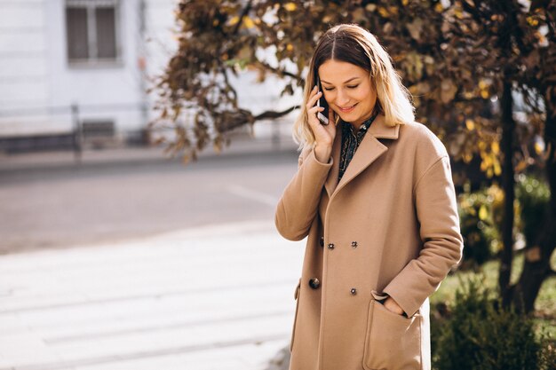 Mujer joven en abrigo beige con teléfono fuera de la calle