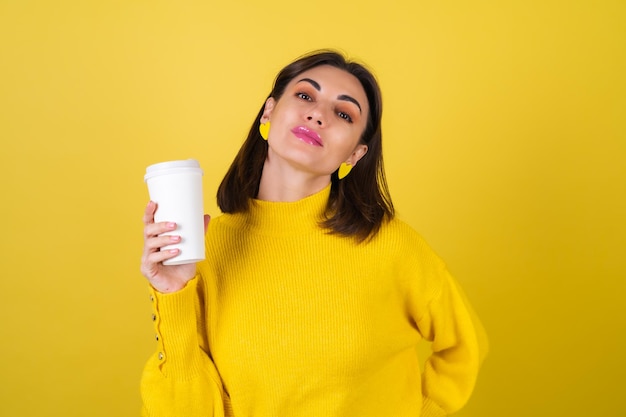 Mujer joven con un acogedor suéter amarillo con brillo de labios rosa brillante con una taza de papel de café aromático caliente, apariencia soñadora, linda sonrisa, irradia calidez