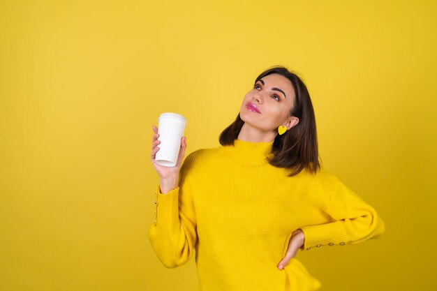 Mujer joven con un acogedor suéter amarillo con brillo de labios rosa brillante con una taza de papel de café aromático caliente, apariencia soñadora, linda sonrisa, irradia calidez