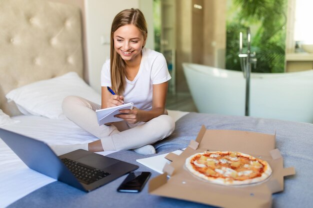 Mujer joven acostada en una cama en el dormitorio en el interior de su casa mientras come pizza y usa la computadora portátil