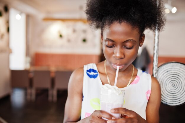 Mujer joven afroamericana pura en vestido de verano en el café bebiendo batido