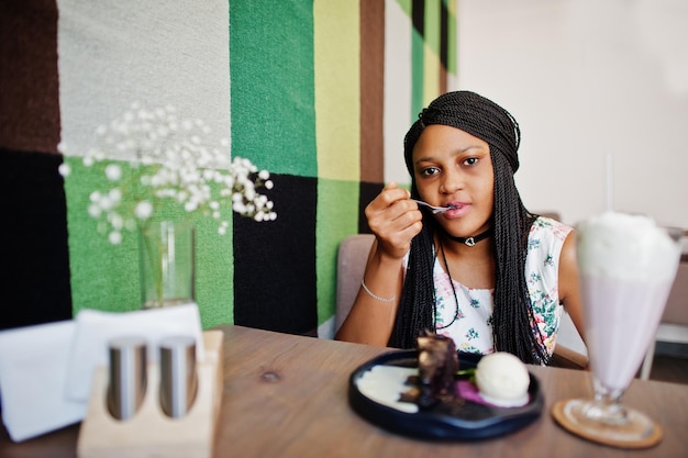 Foto gratuita mujer joven afroamericana pura en vestido de verano y collar de gargantilla en el café comiendo pastel de chocolate con helado