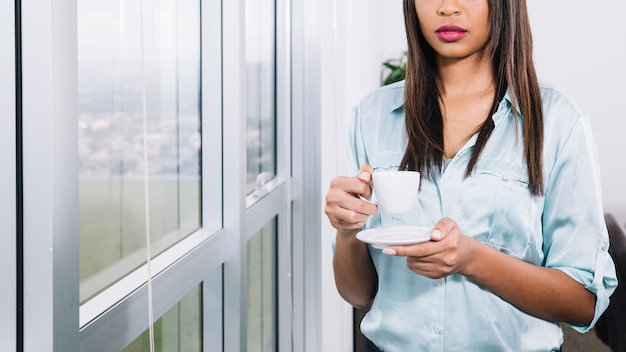 Mujer joven afroamericana con taza junto a la ventana