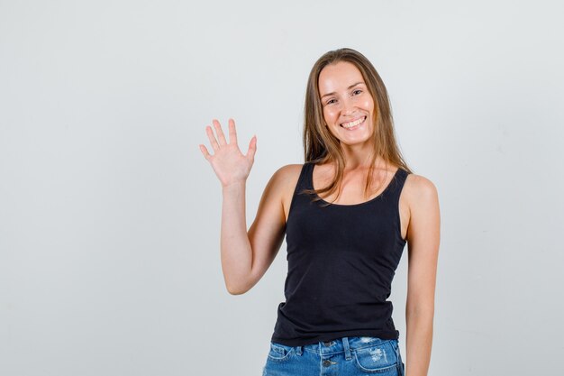 Mujer joven agitando la mano en camiseta, pantalones cortos y mirando alegre. vista frontal.