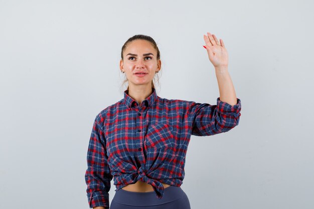 Mujer joven agitando la mano para saludar en camisa a cuadros, pantalones y mirando confiado, vista frontal.