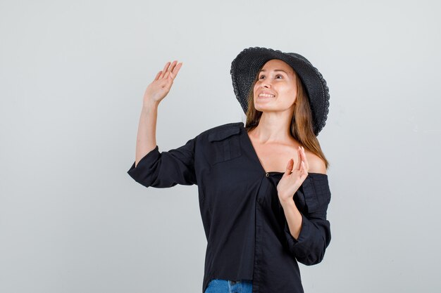 Mujer joven agitando las manos mientras mira hacia arriba en camisa, pantalones cortos, sombrero y mirando alegre