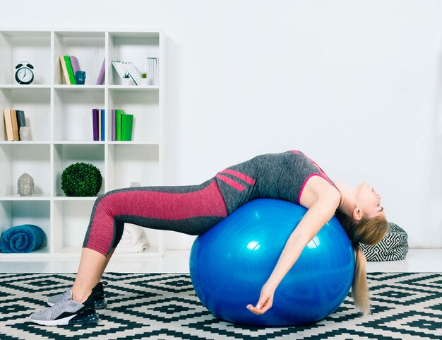 Mujer joven agotada que duerme en la bola azul de los pilates sobre la alfombra