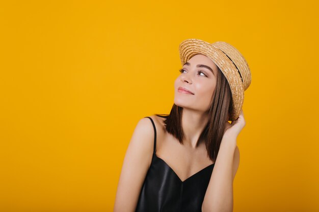 Mujer joven agraciada en la camiseta sin mangas de seda que disfruta. Retrato de niña hermosa tocando su sombrero de paja.