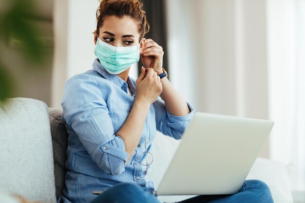 Mujer joven ajustando la máscara facial mientras usa la computadora en casa durante la epidemia de virus