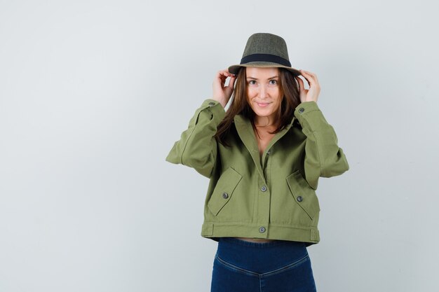 Mujer joven ajustando su sombrero en chaqueta, pantalón y luciendo lindo. vista frontal.