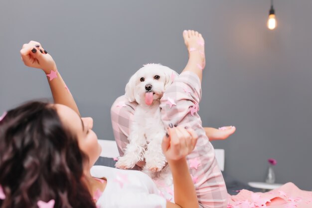 Mujer joven alegre con cabello rizado morena en pijama escalofriante en la cama con perrito en apartamento moderno. Bonita modelo divirtiéndose en casa con mascotas domésticas, expresando felicidad