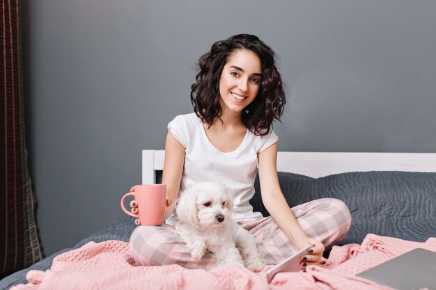 Mujer joven alegre con cabello rizado morena en pijama escalofriante en la cama con perrito en apartamento moderno. Bonita modelo relajándose en casa con una taza de café, charlando por teléfono, sonriendo