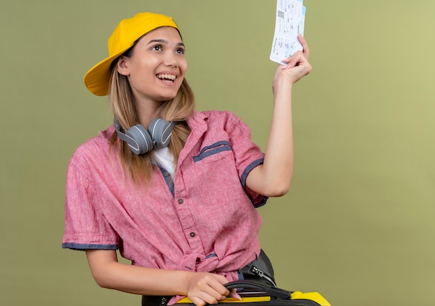 Una mujer joven alegre con camisa roja y gorra de béisbol amarilla que muestra los boletos de avión mientras lleva una maleta amarilla en una pared verde