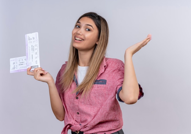 Foto gratuita una mujer joven alegre con camisa roja sosteniendo boletos de avión mientras mira en una pared blanca