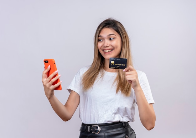 Una mujer joven alegre en camiseta blanca que muestra la tarjeta de crédito mientras mira el teléfono móvil en una pared blanca