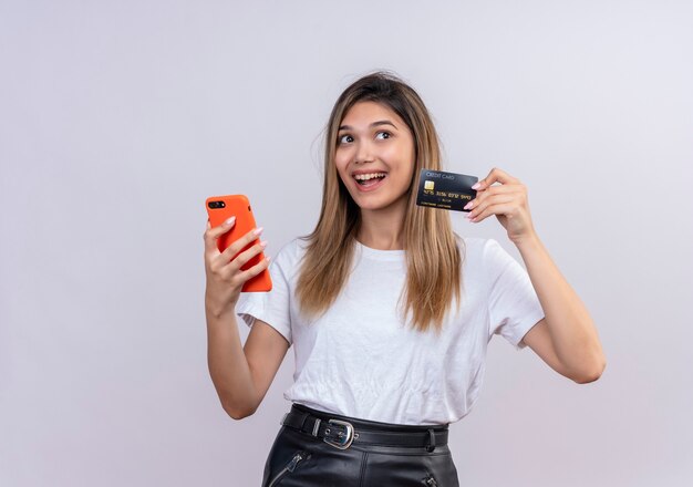 Una mujer joven alegre en camiseta blanca que muestra la tarjeta de crédito mientras sostiene el teléfono móvil en una pared blanca