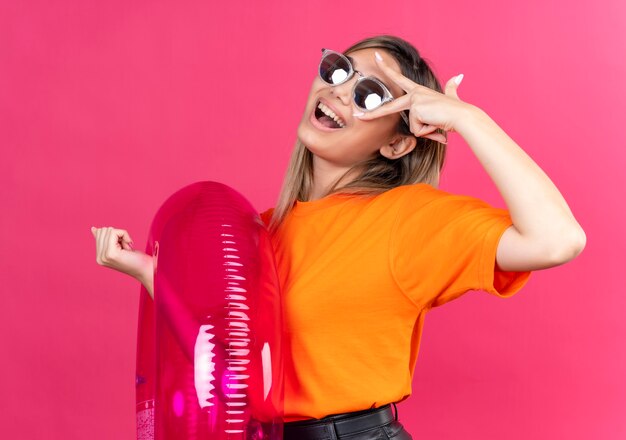 Una mujer joven alegre con una camiseta naranja con gafas de sol sonriendo y mirando mientras sostiene un anillo inflable rosa en una pared rosa