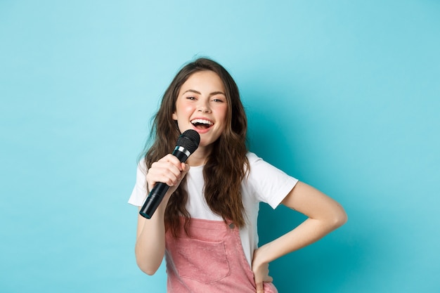 Mujer joven alegre cantando karaoke, sosteniendo el micrófono y sonriendo, divirtiéndose, de pie sobre fondo azul.