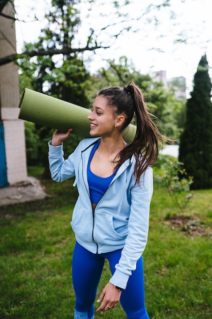 Mujer joven alegre deportes caminando en parque urbano con alfombra de fitness.