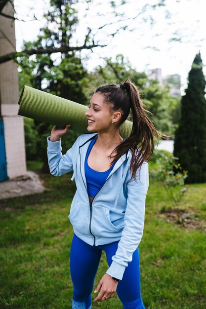 Mujer joven alegre deportes caminando en parque urbano con alfombra de fitness.