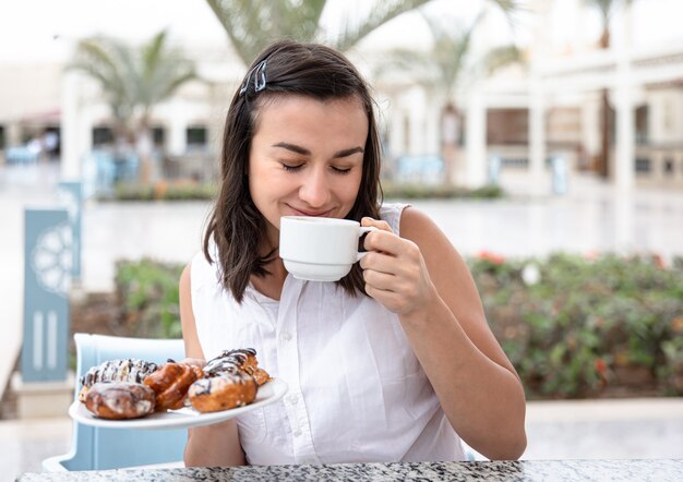 Mujer joven alegre disfrutando de un café por la mañana con donas en la terraza al aire libre