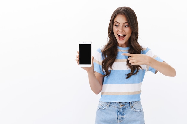 Mujer joven alegre divertida presenta la aplicación de teléfono inteligente, sostiene el teléfono móvil, apunta y mira la pantalla, sonríe emocionada recomienda una aplicación impresionante, tienda en línea