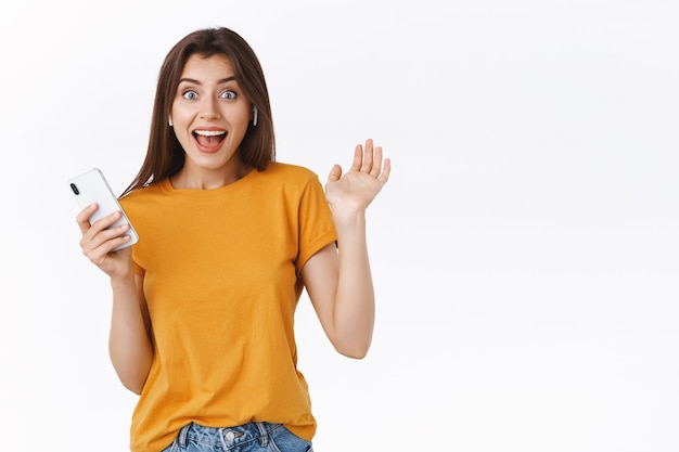 Una mujer joven alegre y emocionada recibe auriculares inalámbricos como regalo de cumpleaños, no puede ocultar la felicidad, se da la mano emocionada y feliz, sostiene un teléfono inteligente, escucha música en auriculares