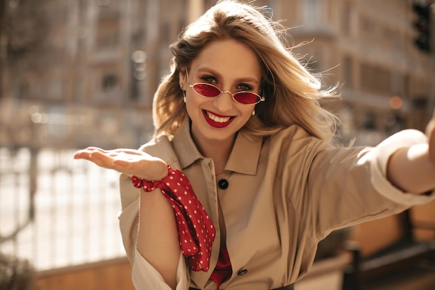 Mujer joven alegre en gabardina beige sonríe sinceramente fuera Feliz mujer rubia con labios rojos toma selfie al aire libre