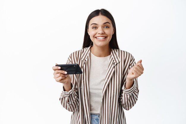 Mujer joven alegre ganando dinero en el teléfono, lograr el objetivo en la aplicación de teléfono inteligente, sosteniendo el teléfono móvil horizontalmente y decir que sí, pared blanca