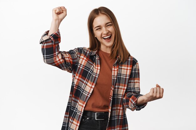 Mujer joven alegre logra el éxito de la meta, grita de alegría, puñetazo, anima al equipo, gana logros, premios, ganando y triunfando, pared blanca