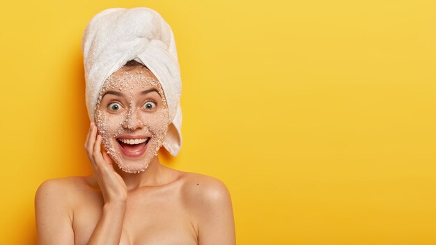 Mujer joven alegre con mascarilla peel off, tiene piel sensible, sonríe ampliamente, muestra dientes blancos, mira feliz, disfruta de frescura en su tez, usa una toalla blanca en la cabeza, modelos de interior sin camisa