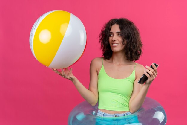 Una mujer joven alegre con el pelo corto en la parte superior verde de la cosecha que mira la bola inflable que sostiene el teléfono móvil