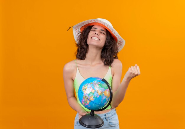Foto gratuita una mujer joven alegre con el pelo corto en verde crop top vistiendo sombrero para el sol sosteniendo un globo con puño cerrado