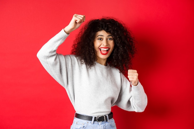 Mujer joven alegre con el pelo rizado, levantando la mano y celebrando la victoria, lograr la meta o el éxito, de pie sobre fondo rojo.