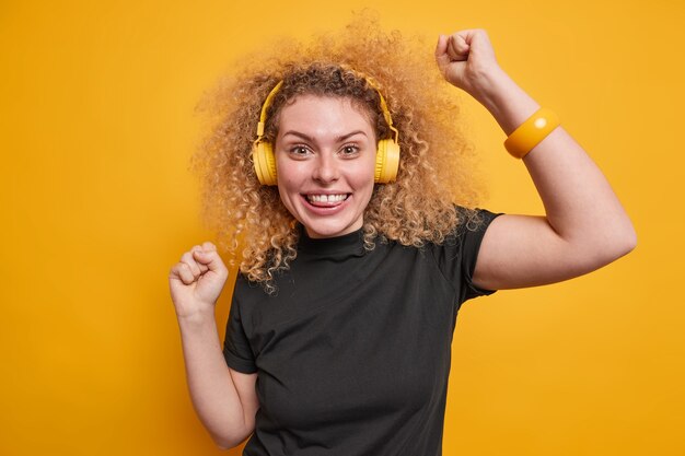 La mujer joven alegre con el pelo rizado natural levanta los brazos baila sin preocupaciones disfruta de su música favorita en los auriculares se divierte vestida con una camiseta negra aislada sobre una pared amarilla. Concepto de entretenimiento