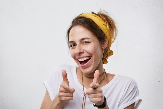 Mujer joven alegre positiva con pañuelo amarillo en la cabeza y camiseta blanca casual parpadeando y sonriendo señalando con los dedos índices. Feliz mujer atractiva apuntando a ti