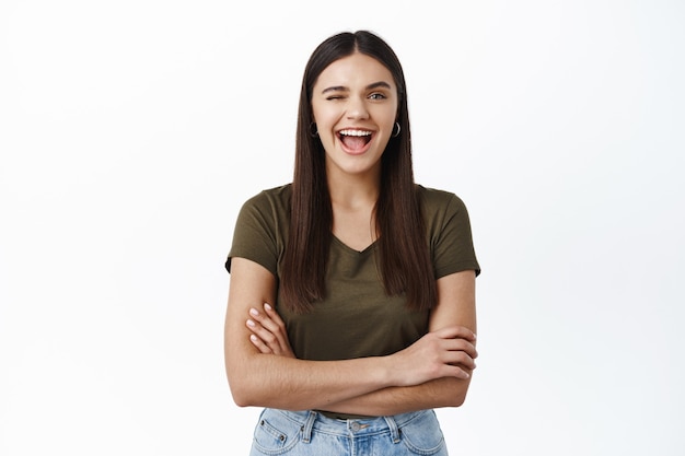 Mujer joven alegre y positiva que te anima, guiñando un ojo y sonriendo afirmativamente, insinuando que todo sea bueno, de pie contra la pared blanca con los brazos cruzados