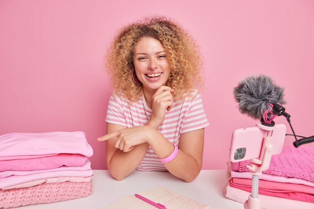 Foto gratuita mujer joven alegre con puntos de pelo rizado en la pila de ropa limpia doblada da consejos sobre la limpieza para sus seguidores toma notas en el bloc de notas se sienta en la mesa frente a la cámara del teléfono inteligente